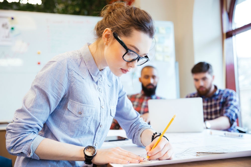 Image of a woman drafting with a pencil. Two people sit in the background working on a computer.