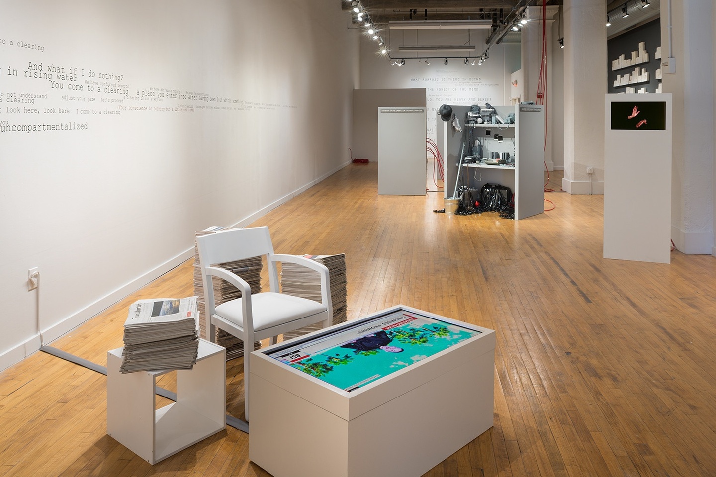 Installation view of a gallery space: in the foreground, a chair next to a video on a screen in a white platform/box, with stacks of newspapers surrounding this all. In the background, wires from exposed machinery and other artworks mounted to the wall. The walls are white with some text running along it in a typewriter font. The floors are wooden.