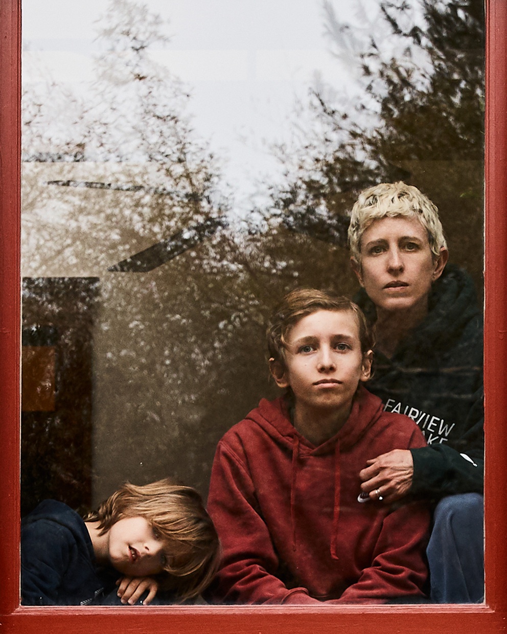 A photograph of a red framed window. A light-skinned mother and two light-skinned children look out at the viewer from behind the window pane.