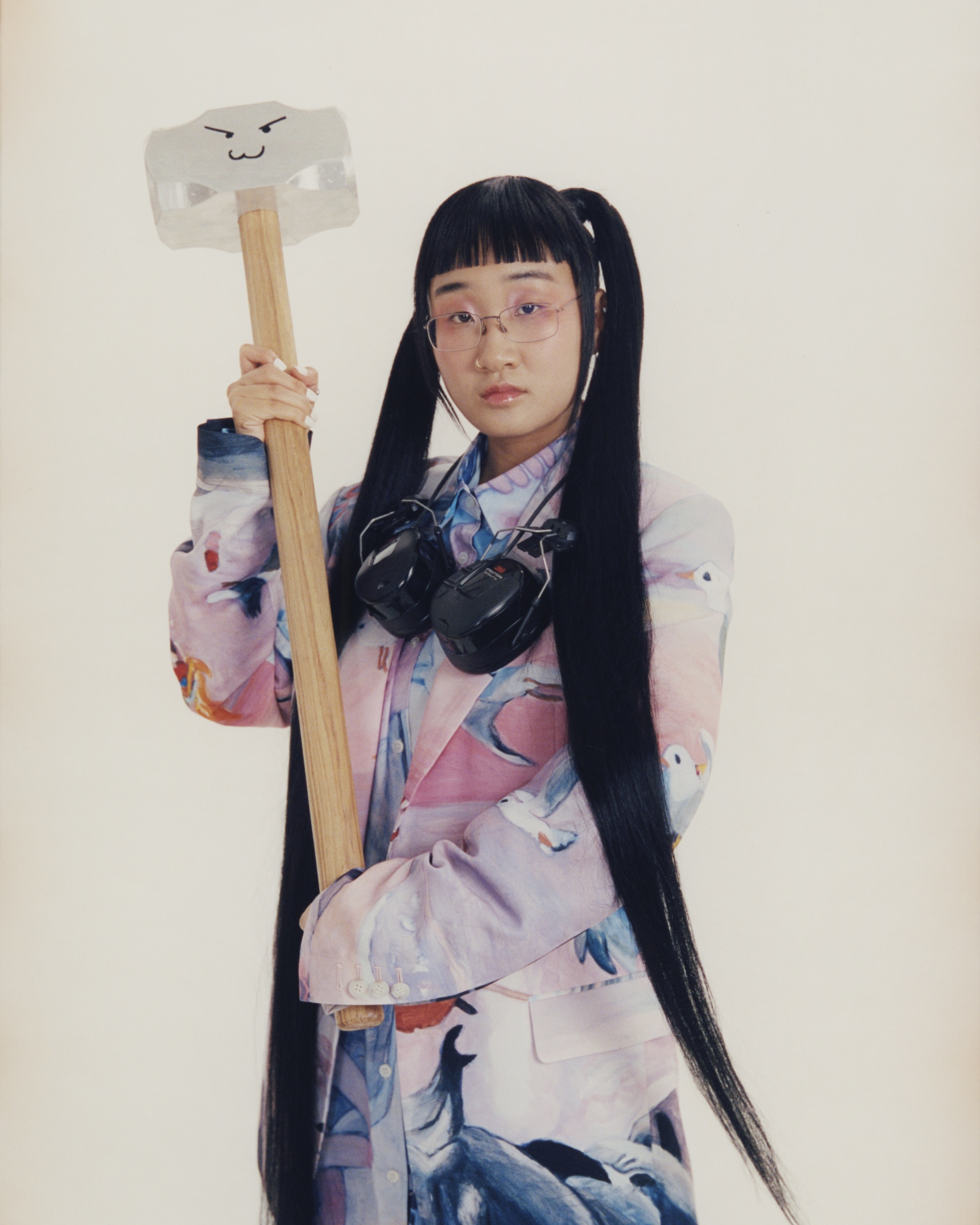 A Korean American woman with long, dark hair sweeping down beneath her hips holds up a large sledgehammer and looks directly at us. 