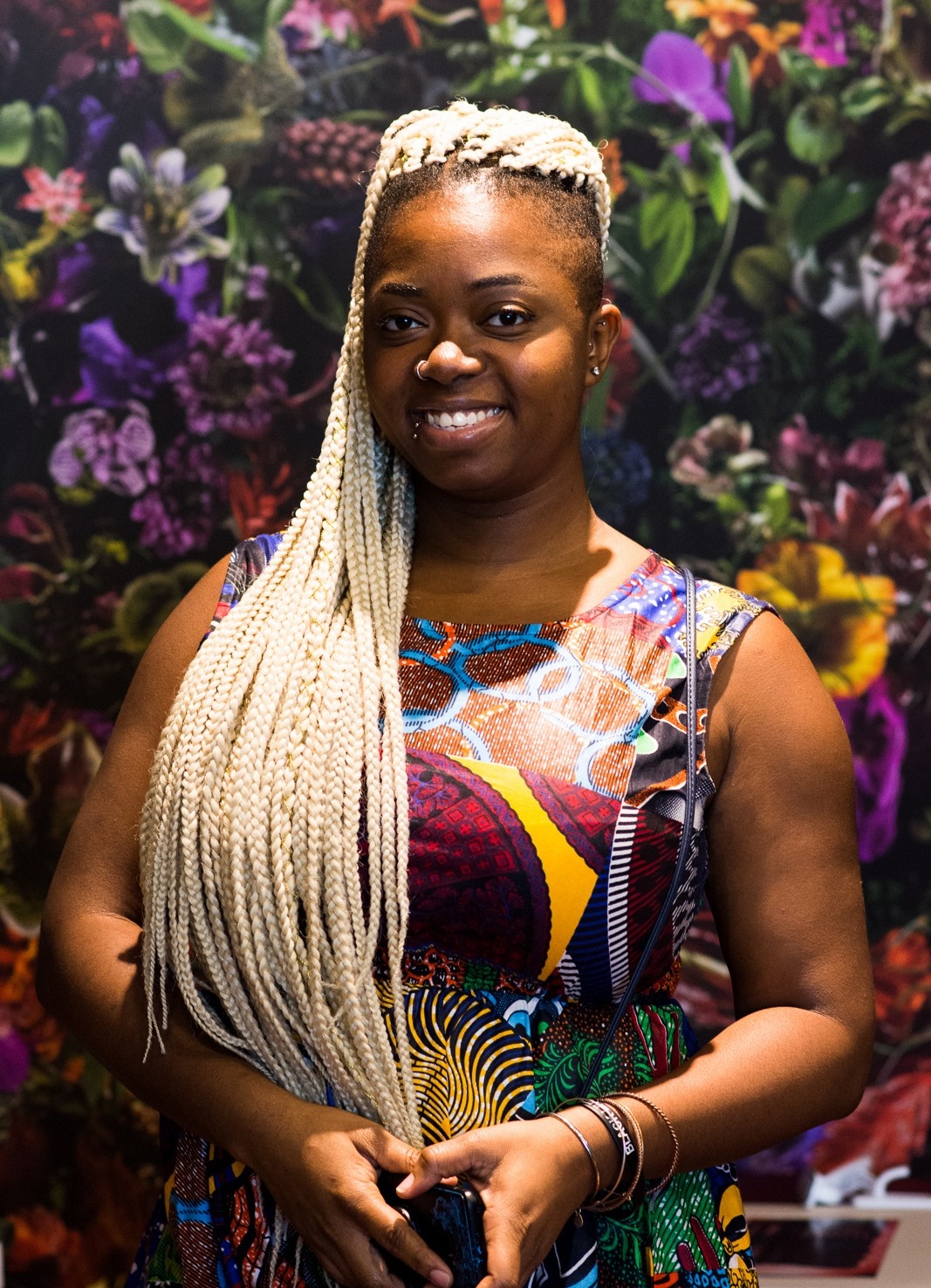 The artist Tasha Dougé smiling in a dress with a bright print against a floral background. Her hair is in braids down her shoulder and chest, and her hands are together in front of her waist.