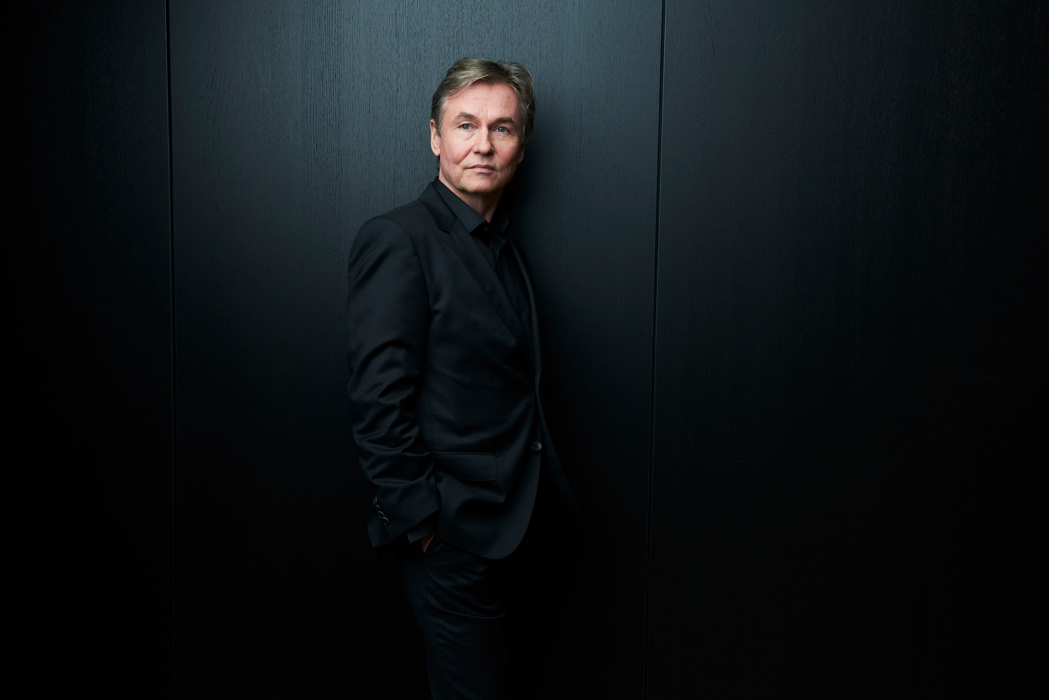 Conductor Esa-Pekka Salonen standing against a black background. He wears a black suit that blends into the shadowy background and his face and head are dramatically lit by a focused light.