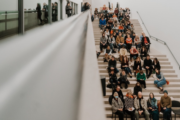 View of crowd seated on 3 tiers of steps facing the panelists. 