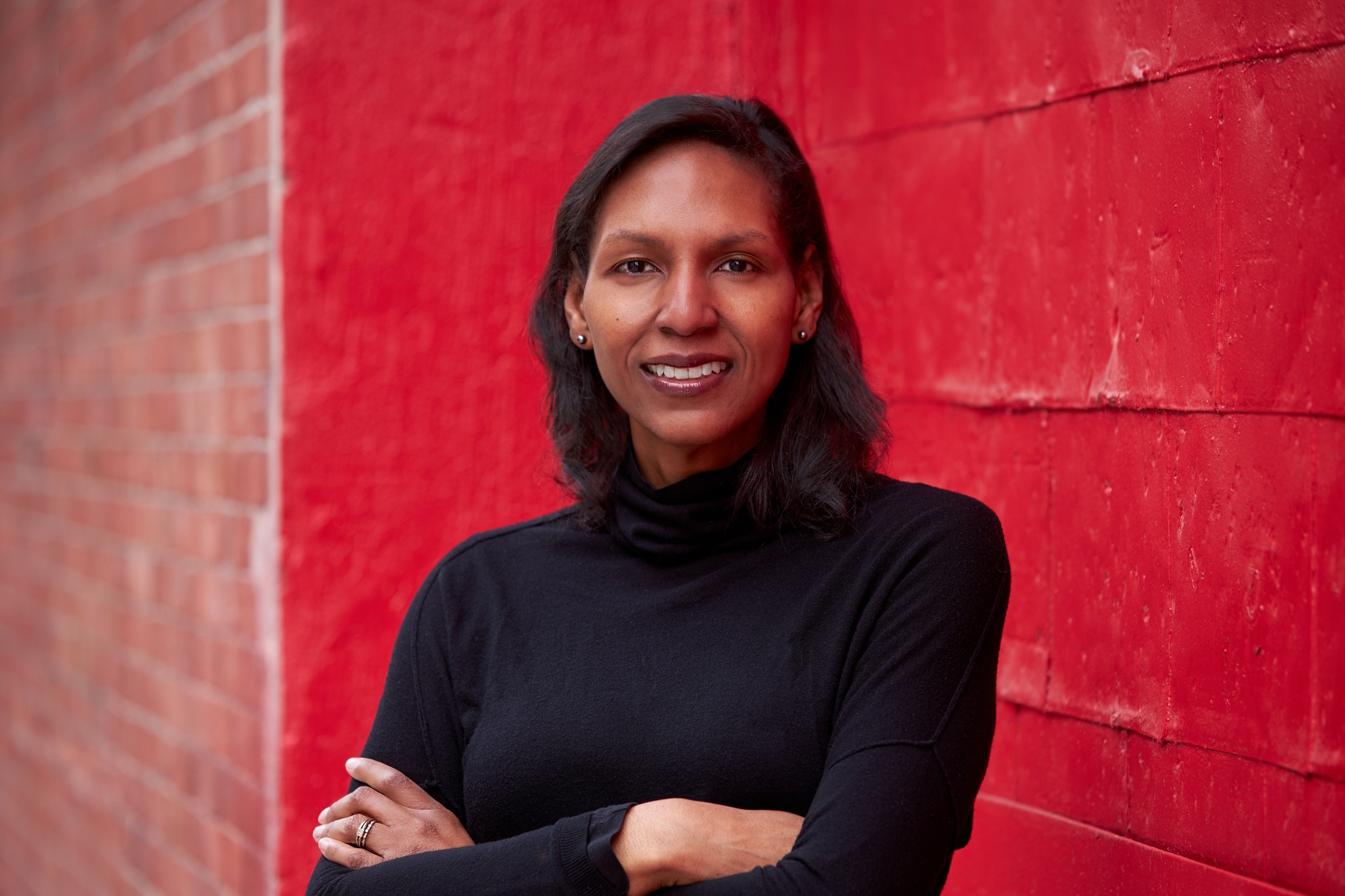 A photo of curator, art historian, and professor Courtney J. Martin wearing a black long-sleeve turtle neck and smiling with arms crossed against a wall painted a bright red. 