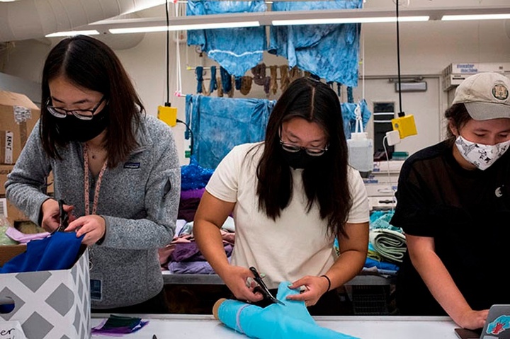 Three students wearing COVID masks working side-by-side in the fashion design studio, with various fabrics out.