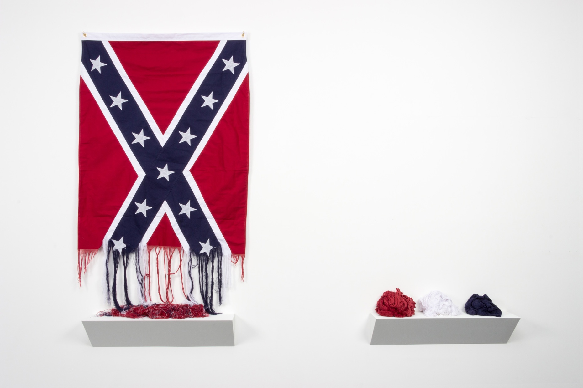 An unraveling Confederate flag hung on a white wall next to a shelf with balls of red, white, and blue fabric.