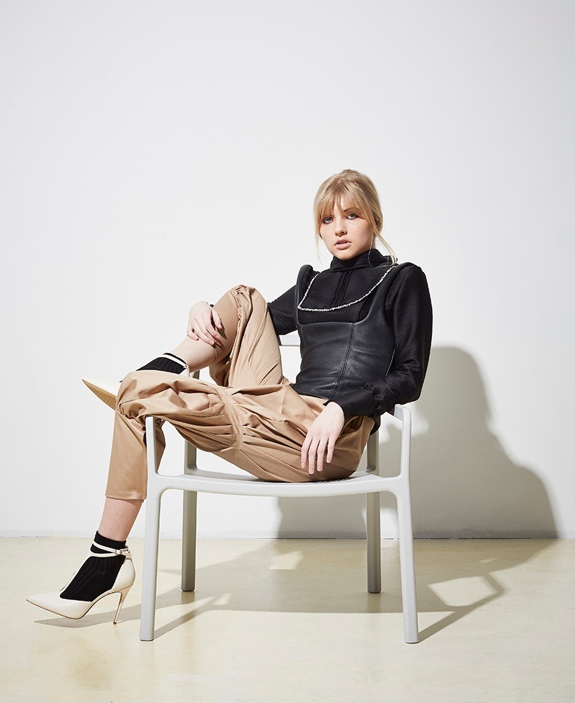 Model lounges in a chair wearing a a black turtleneck with rhinestone detailing, a black leather corset top, and khaki jodhpur style trousers.