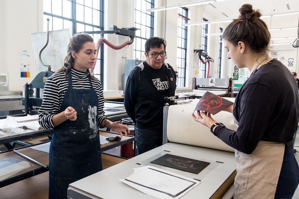 Person checks the quality of a print of a black wolfish head on red paper while two others observe.