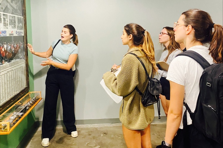 One person talking and gesturing at framed poster on wall while three students stand and listen.