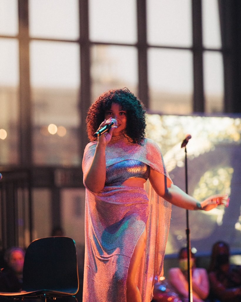 A Black woman with curly hair to her shoulders speaks into a microphone during a performance. Behind her are tall windows with dusky sunlight and a video screen and members of the audience.