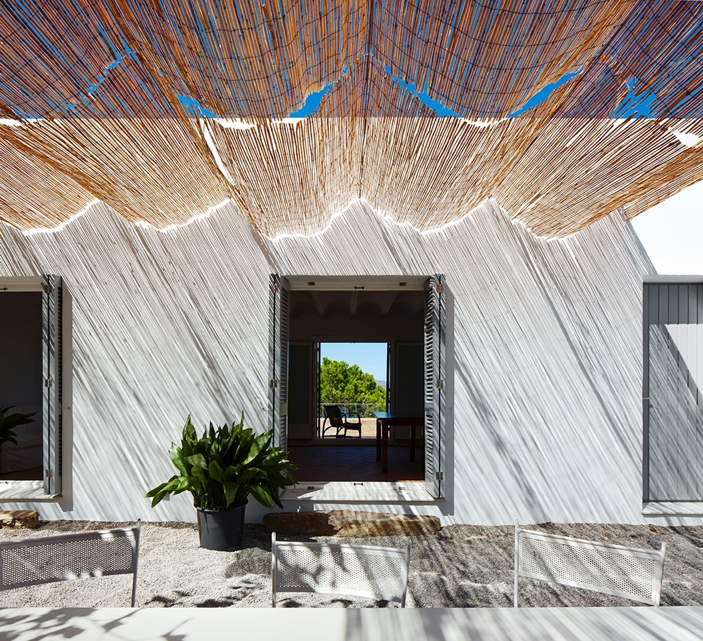 A white house with bamboo awning that throws lined shadows onto the scene. The double doors are opened and we can see through the house and out the other opened double doors on the other side