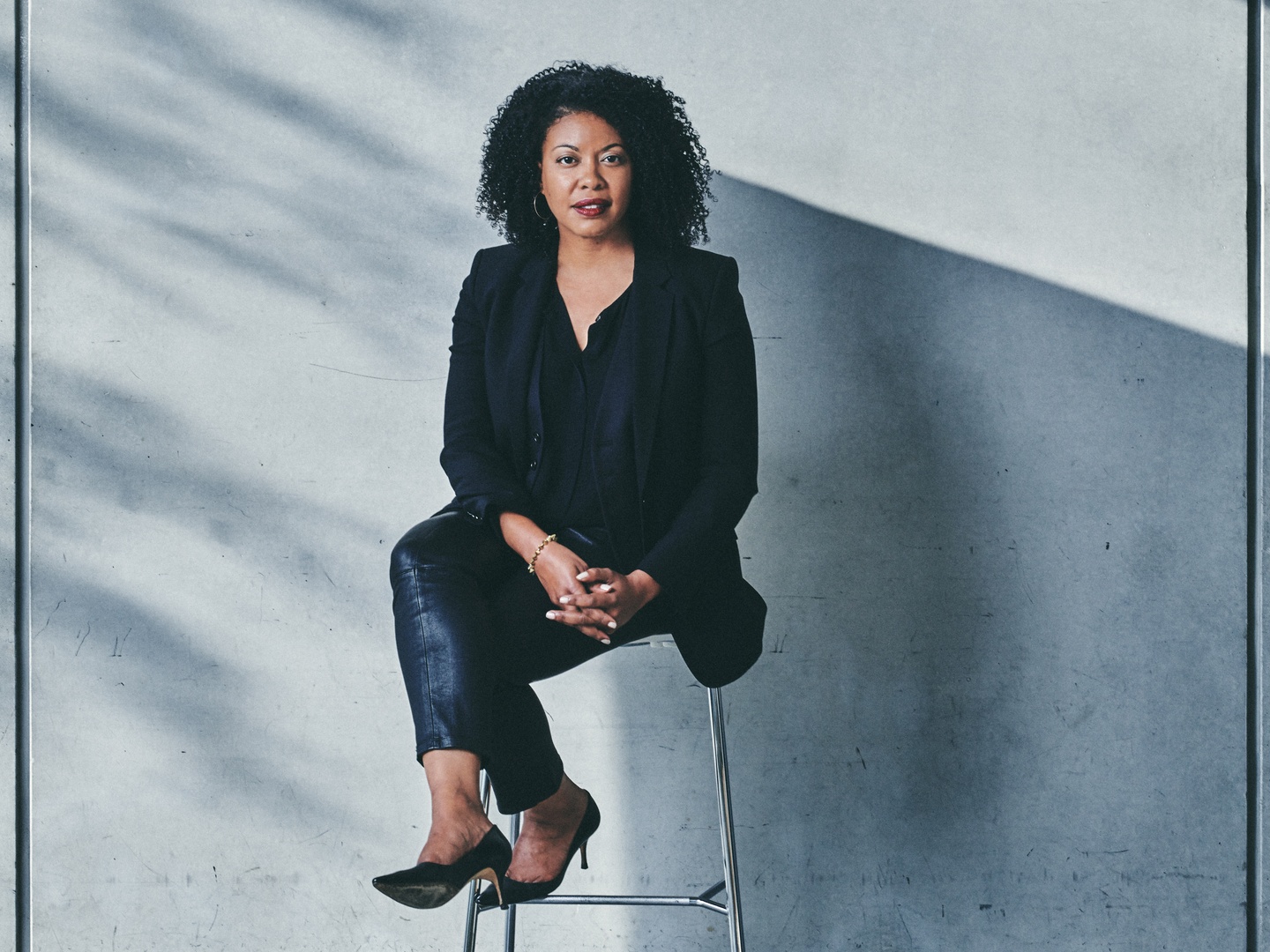 A photo of a woman dressed in black seated in front of a gray geometric backdrop