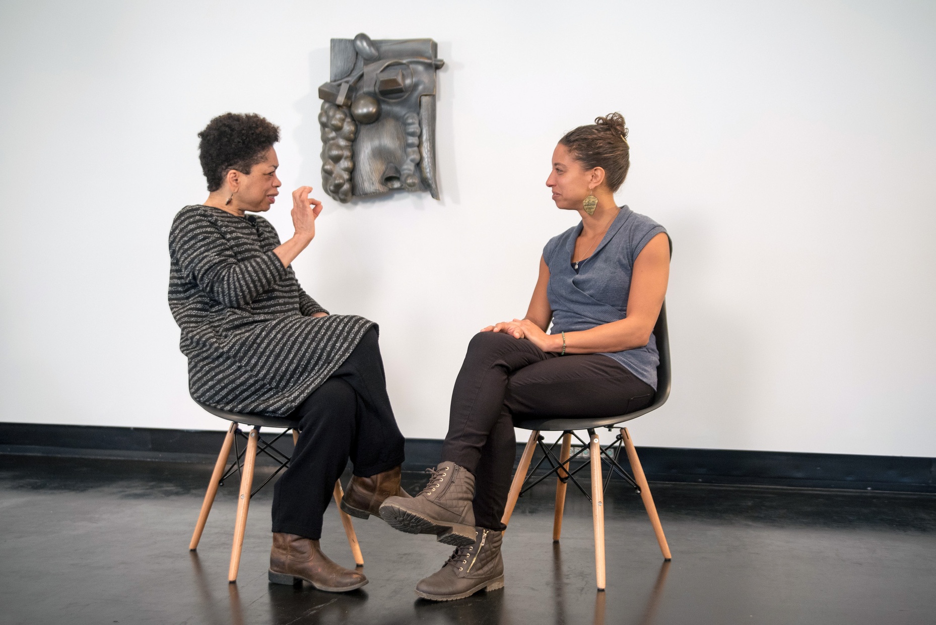 Syd Carpenter sits on a chair in the left side of the photo looking towards Leah Penniman, angled towards Syd in a chair in the right side of the photograph. Syd's sculpture is mounted on the white wall behind them and is visible between the two figures.