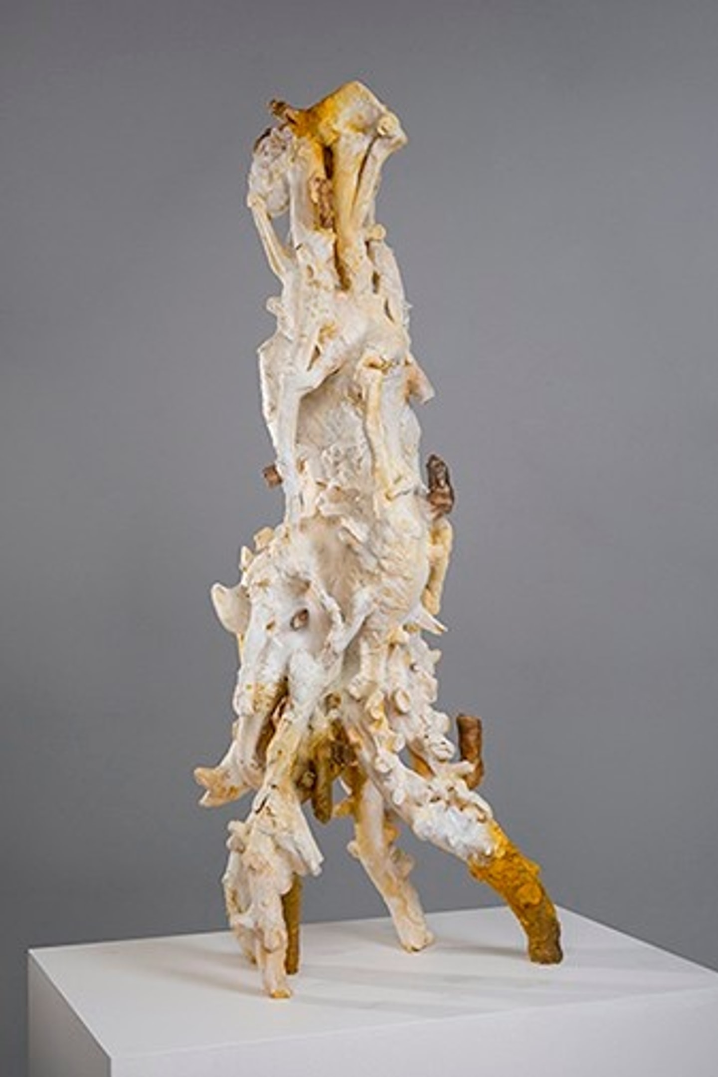 An abstract, tall vertical ceramic sculpture by Arny Nadler featuring fused, bone-like organic forms in ivory and mustard yellow, standing on a white pedestal against a solid grey background.