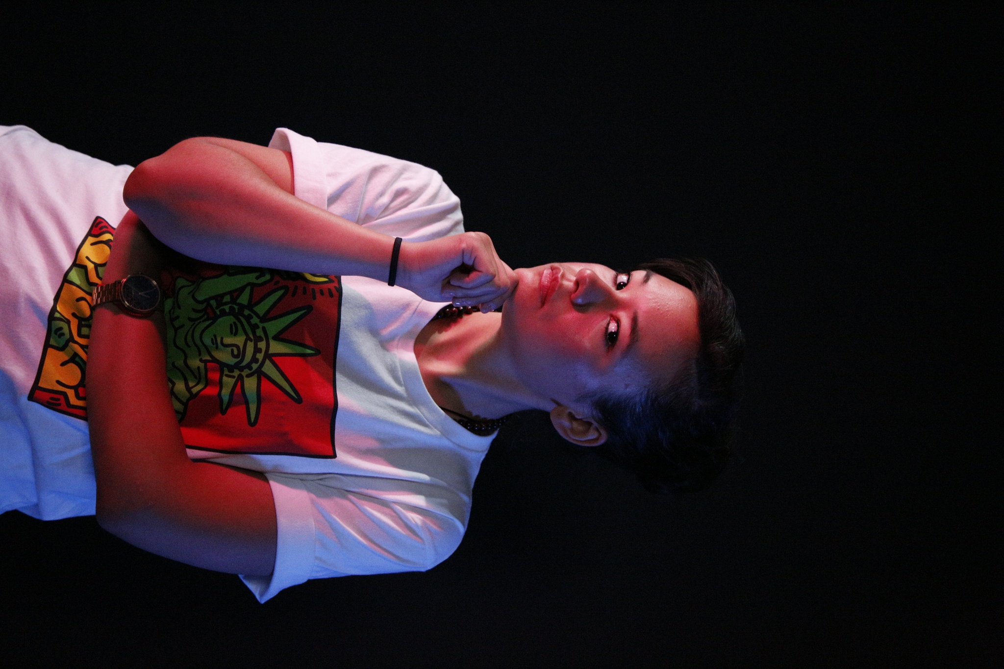 A portrait of Puerto Rican DJ Perly. She has short brown hair parted toward the right side of her head and looks at us sideways at an angle. She holds one hand up under her chin pensively, and wears a white t-shirt with an illustration of the Statue of Liberty.