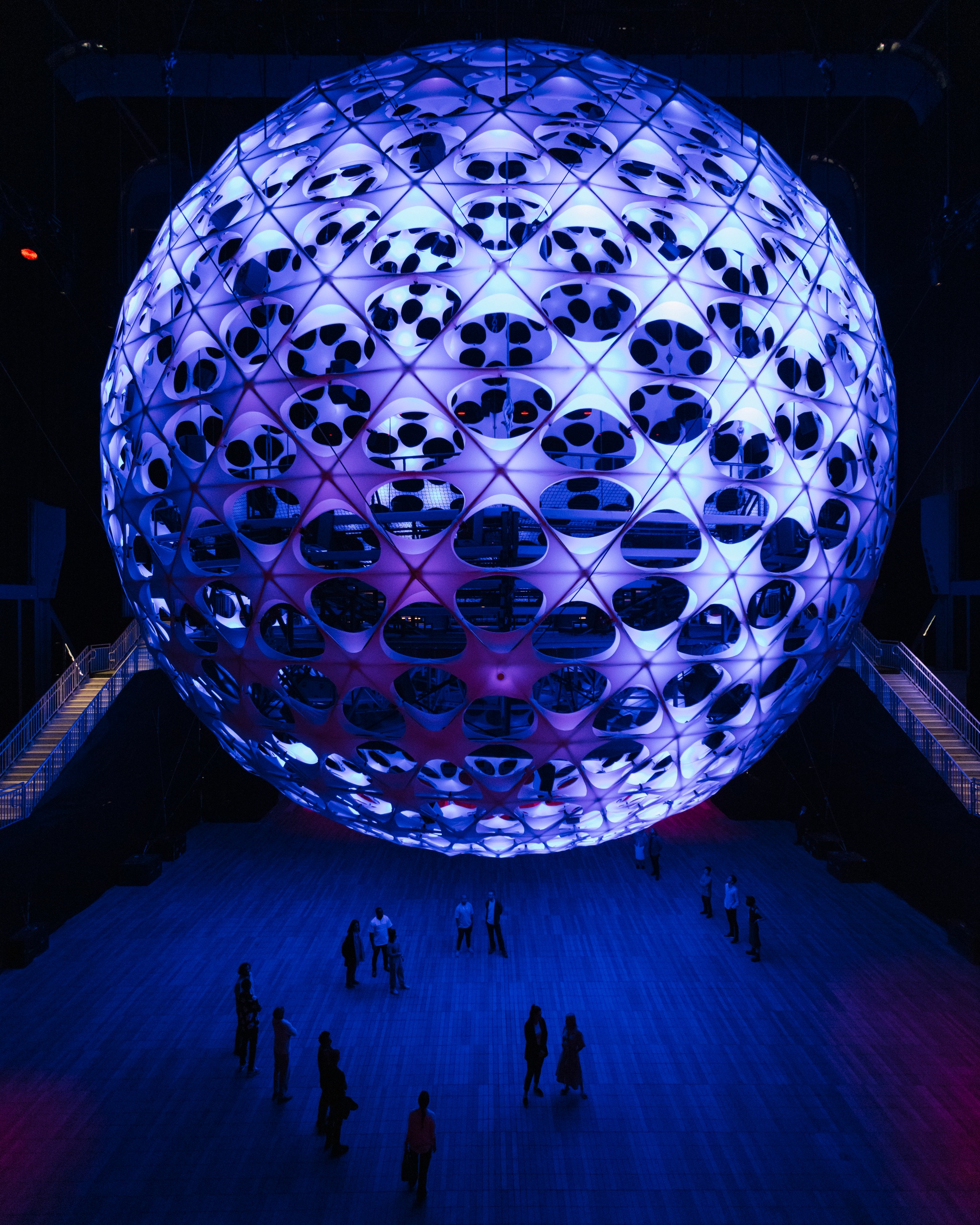 A large spherical structure hangs from a 115-foot-tall ceiling above roughly 20 people who stand below it. The sphere is dotted with light nodes that emanate a purplish blue light. The people are small figures beneath the enormous sphere.