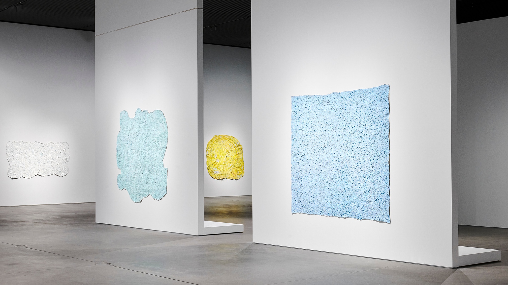Abstract paintings in the exhibition "Howardena Pindell: Rope/Fire/Water." In the foreground are two blue paintings, with a curvilinear yellow painting visible through a gap in the wall in the background, and a white rectangular canvas to the left.