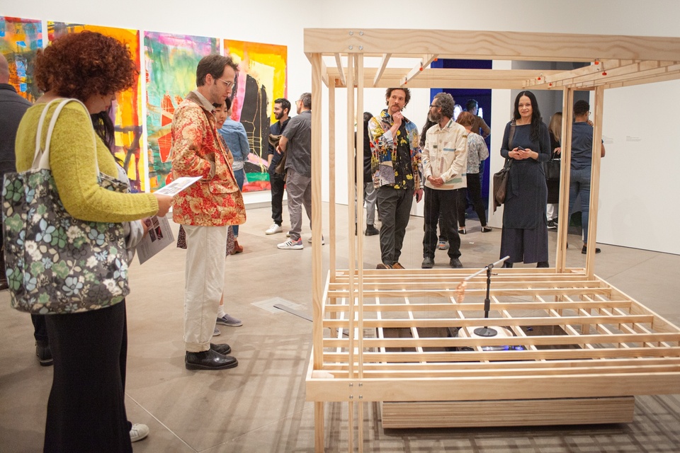 Gallery visitors investigate a wooden construction the size of a small garden shed. A knee-height slatted platform covers a mechanism, which sends a metal pole arm spinning around the platform. The end of the pole has a fan of copper wires that brush against vertical wires strung around the framework.