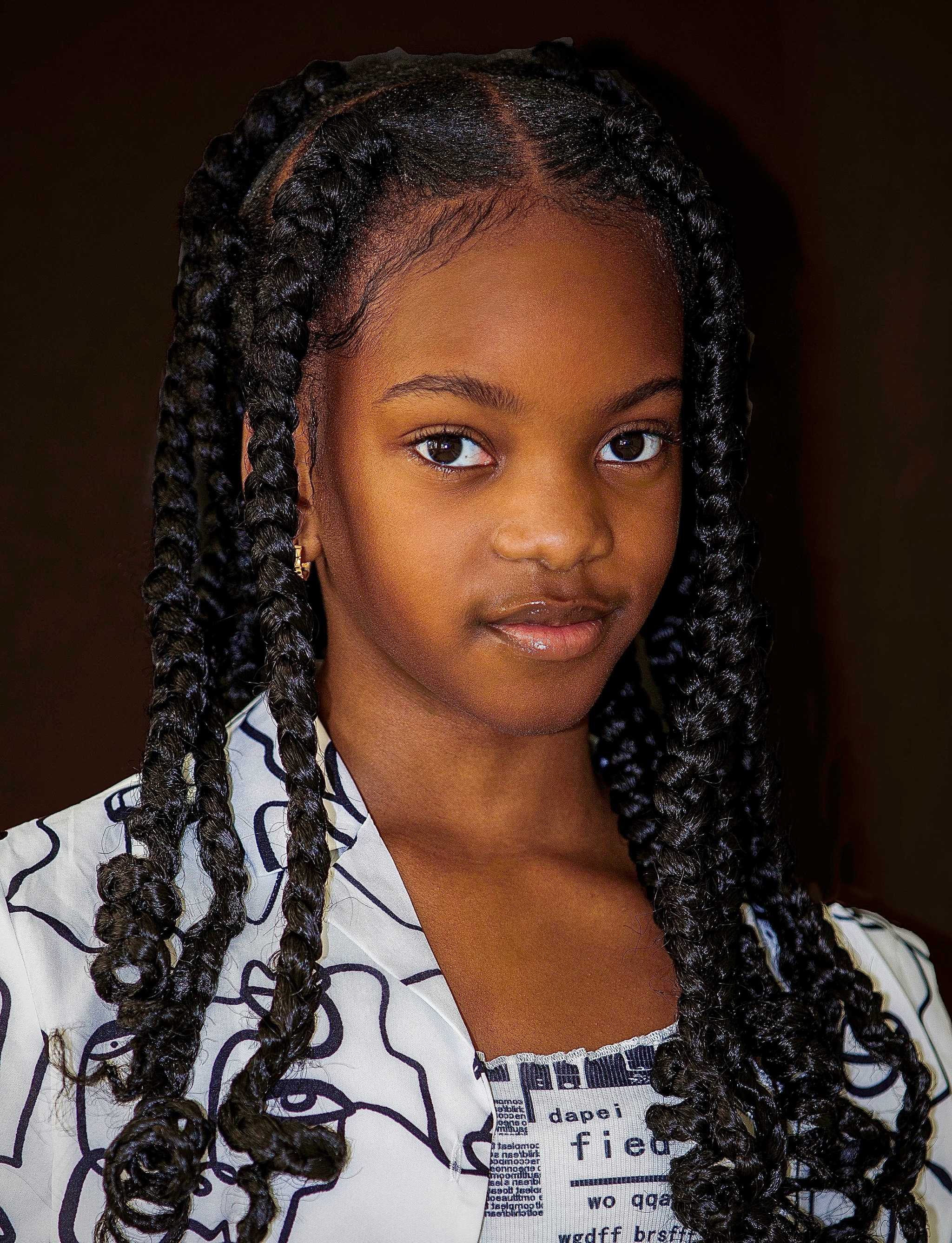 A headshot of child actor Ifeoluwa Adeniyi, young Black girl whose hair is parted down the middle with long locs falling down either side of her head past her shoulders. She wears a white shirt with a black graphic print.