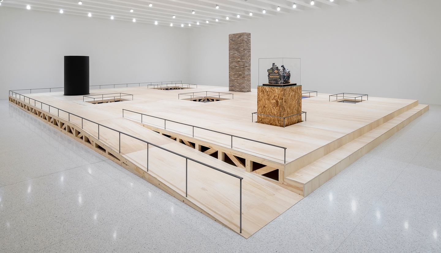 A wood platform with a long ramp in the middle of a white gallery; the platform has various sculptures and voids on the surface