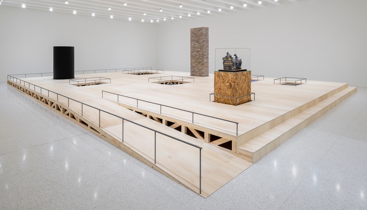A wood platform with a long ramp in the middle of a white gallery; the platform has various sculptures and voids on the surface