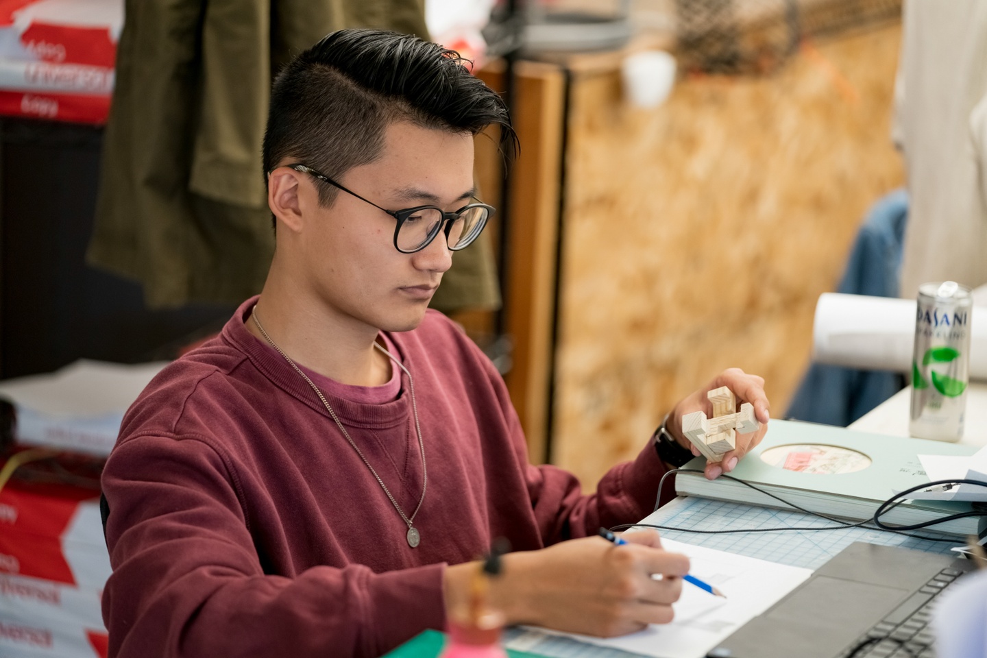 Person sitting at a desk holds a small wooden model in one hand and sketches something with the other.