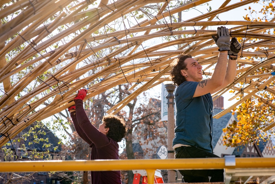 Two people wearing work gloves sit on a scaffold and affix ties to a domed bamboo structure above their heads.