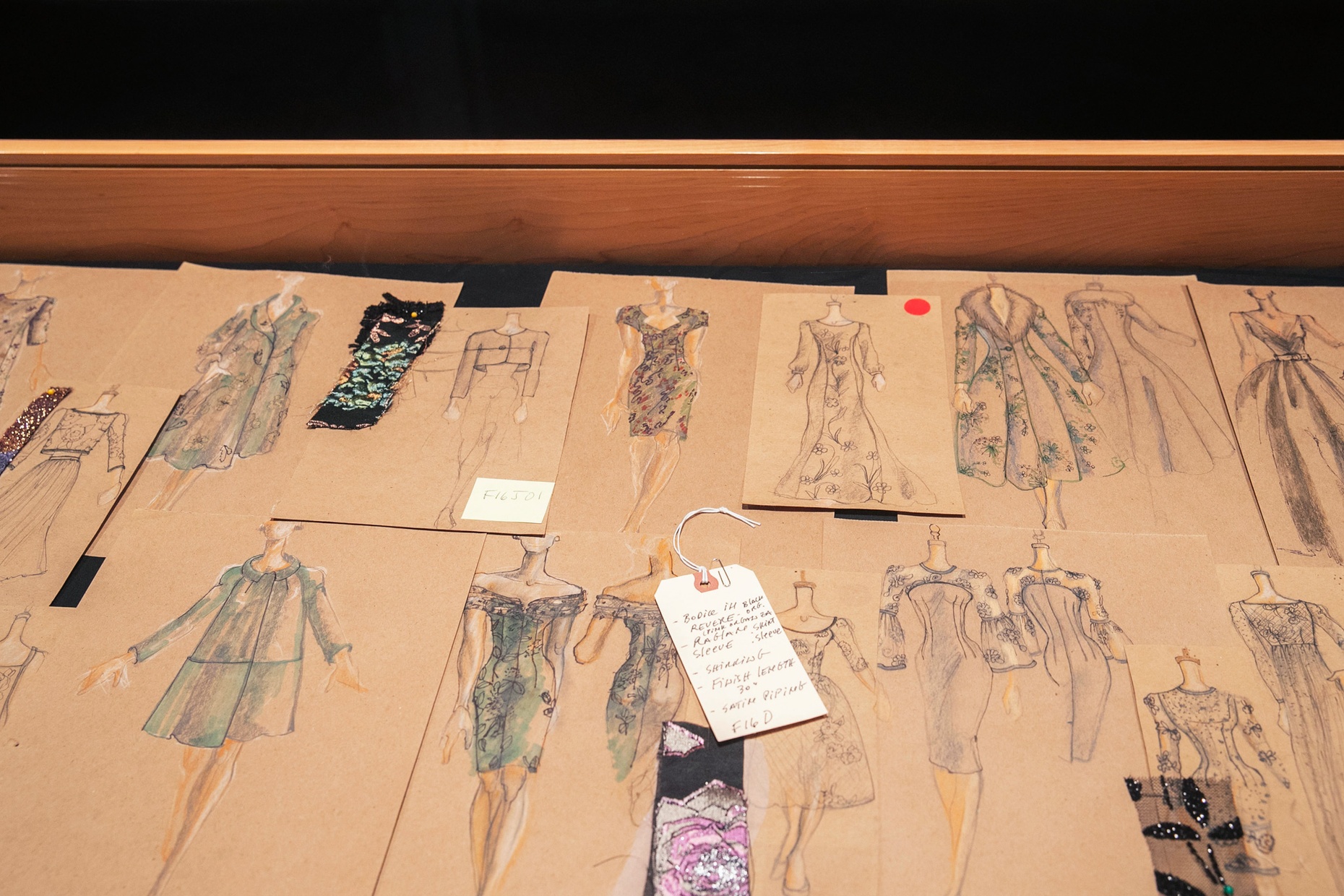 A case filled with fashion drawings, each on brown paper, some with fabrics attached.