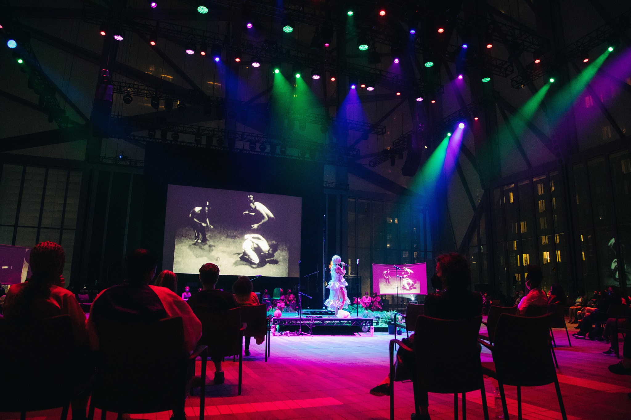 A performer stands on a round platform in the center of an audience, lit with purple and green spotlights. In the background, two video screens project a film of three people dancing.