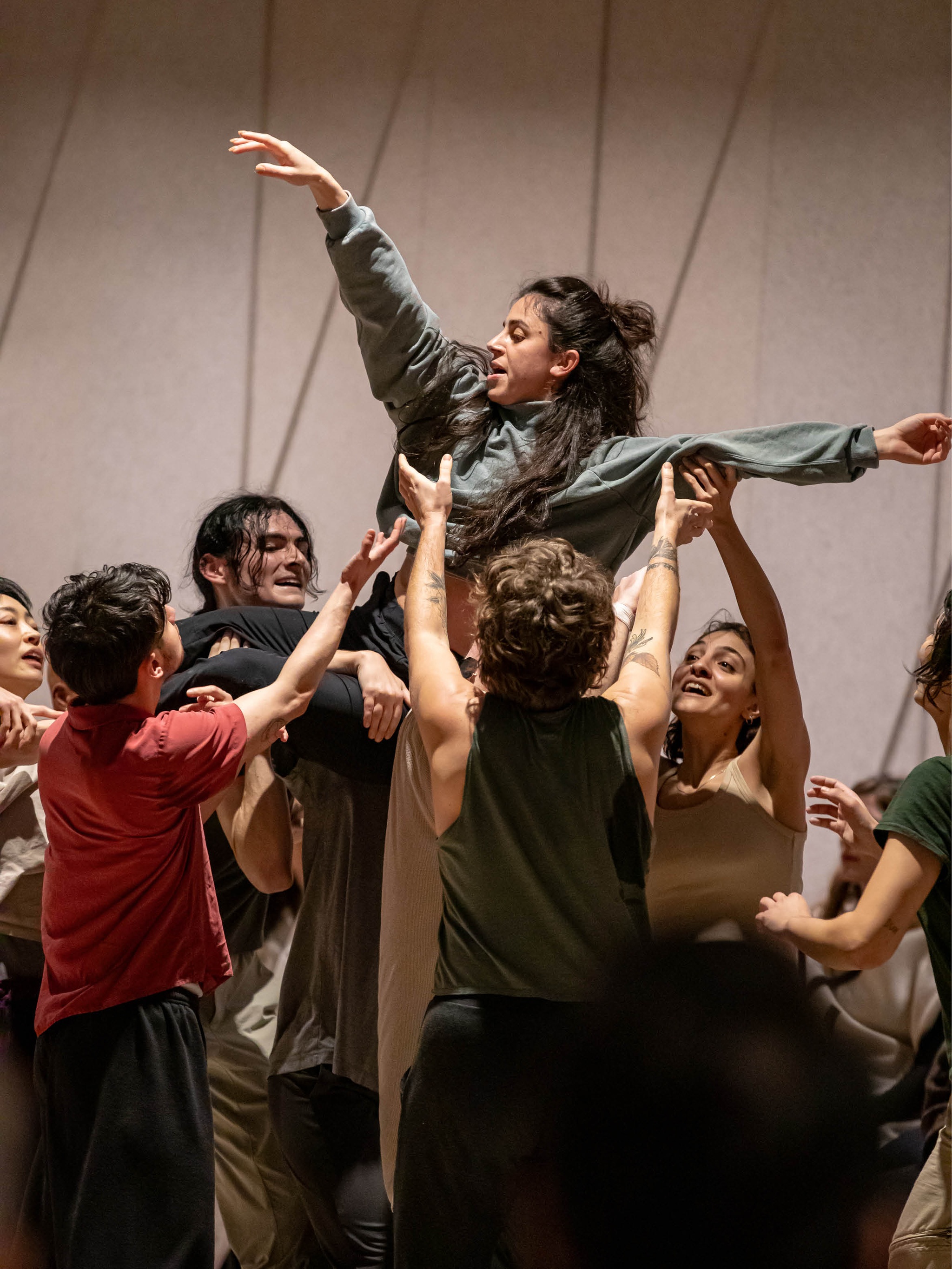 An ensemble of dancers lift another performer overhead. The dancer being held aloft wears a green sweatshirt and gray sweatpants with her arms outstretched exuberantly. She is smiling while the other performers support her.