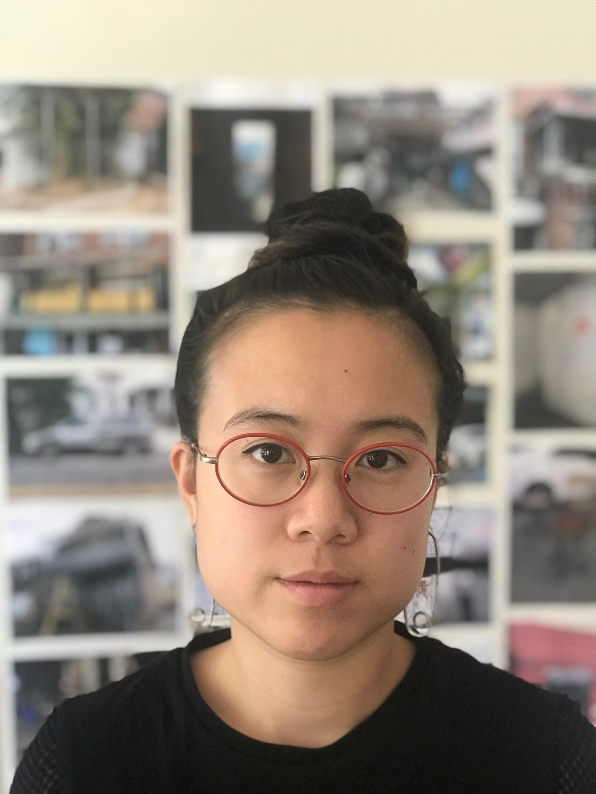 A photo of the artist Anne Wu. She wears red-rimmed glasses with her hair in a bun. In the slightly out-of-focus background, photographs and images are hanging on the wall. 