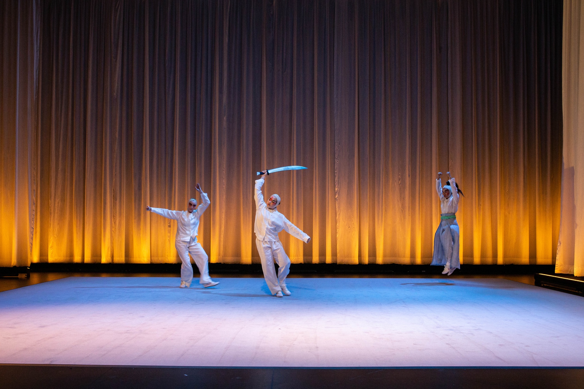 Three dancers in stand across a wide stage lit with a soft purple light. They are each mid gesture with arms extended out and above their heads. At the center, a dancer holds a sword above her head. In the background, a curtain is lit with a warm golden light.
