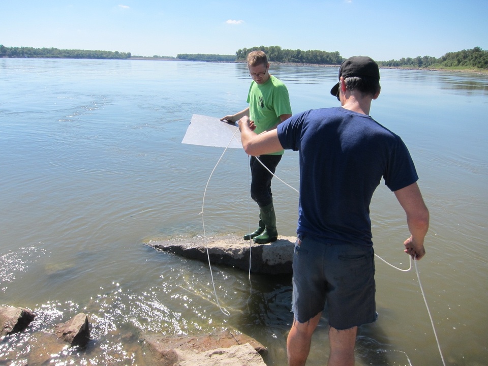 Two people standing precariously on a rock over a body of water holding a piece of string between them