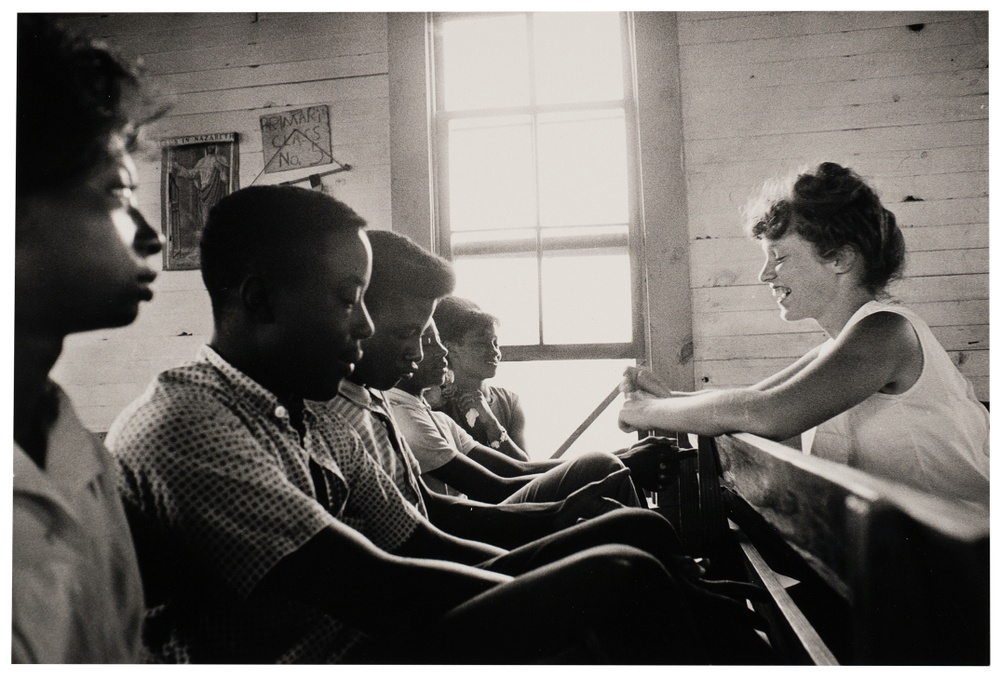 A black and white photograph of five people seated on a bench next to a sun filled window facing a smiling woman.