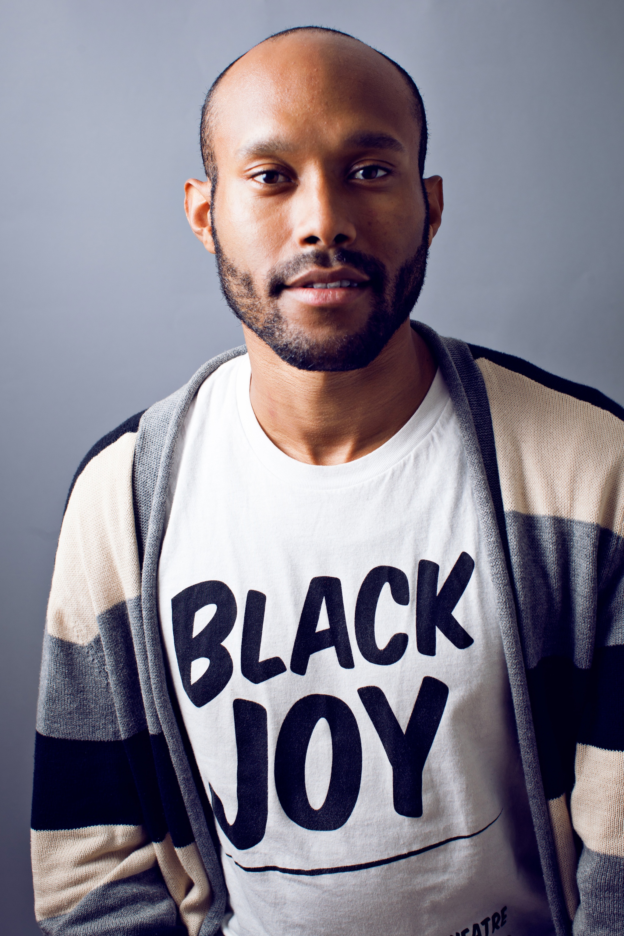 A portrait of Jonathan McCrory, Black man with a scruffy beard and mustache who poses facing us with a tshirt on that reads Black Joy. 