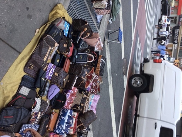 Photo of an artwork installation, oriented on its side. The photo shows a huge pile of handbags of various colors and sizes on a yellow blanket next to a busy street.