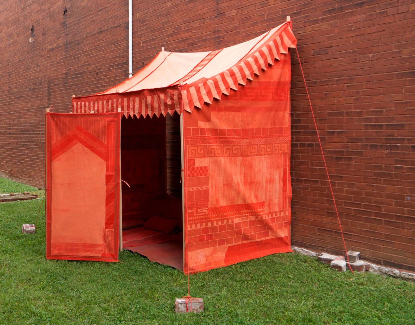 Against a brick wall on green grass, an installation: a tent anchored to the ground with bricks. The tent is bright red/vermilion and light coral, its door open. The interior of the tent is dark. Faint geometric patterns appear on the side of the tent.