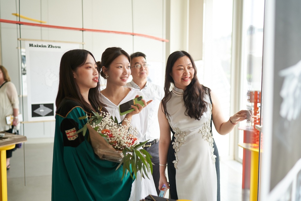 A family with their graduate stands next to an architectural model and reads one of the hanging posters. 