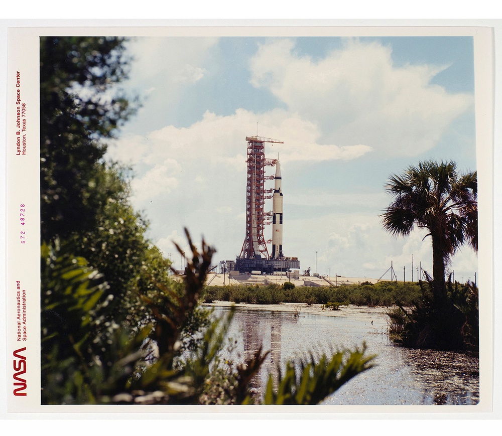 Photograph of a rocket on a launch pad in the distance with shrubbery and swampy water in the foreground.