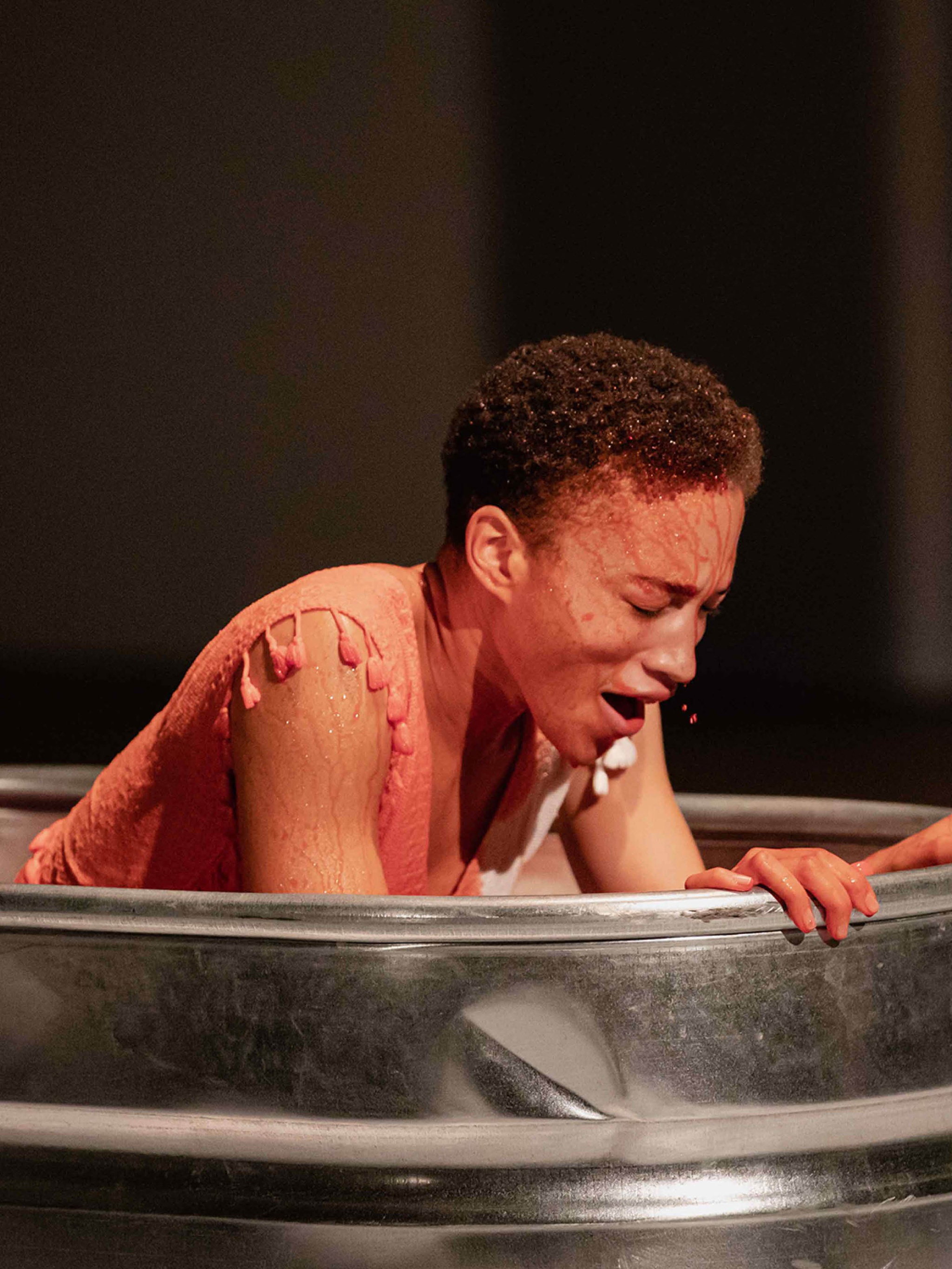 The artist Asia Stewart rises from the red water inside a metal wash tub, gasping for breath with eyes closed. The red liquid pearls in her hair and runs in rivulets down her face, dying the sleeveless white dress she wears red.