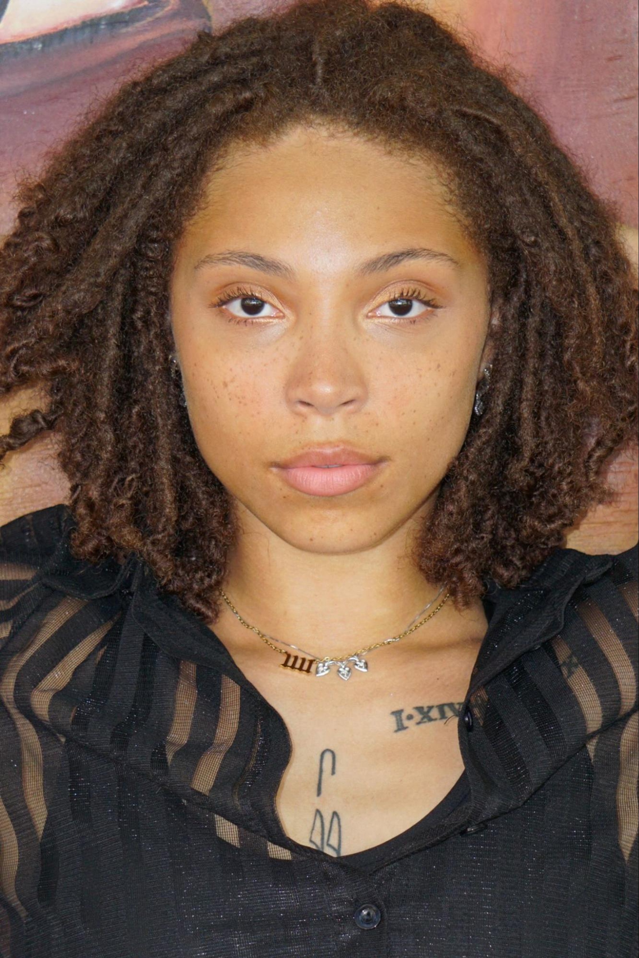 A portrait of Symone Denwood, a dancer with light brown skin and reddish brown locs. She looks directly at us with the hint of a smile on her face. 