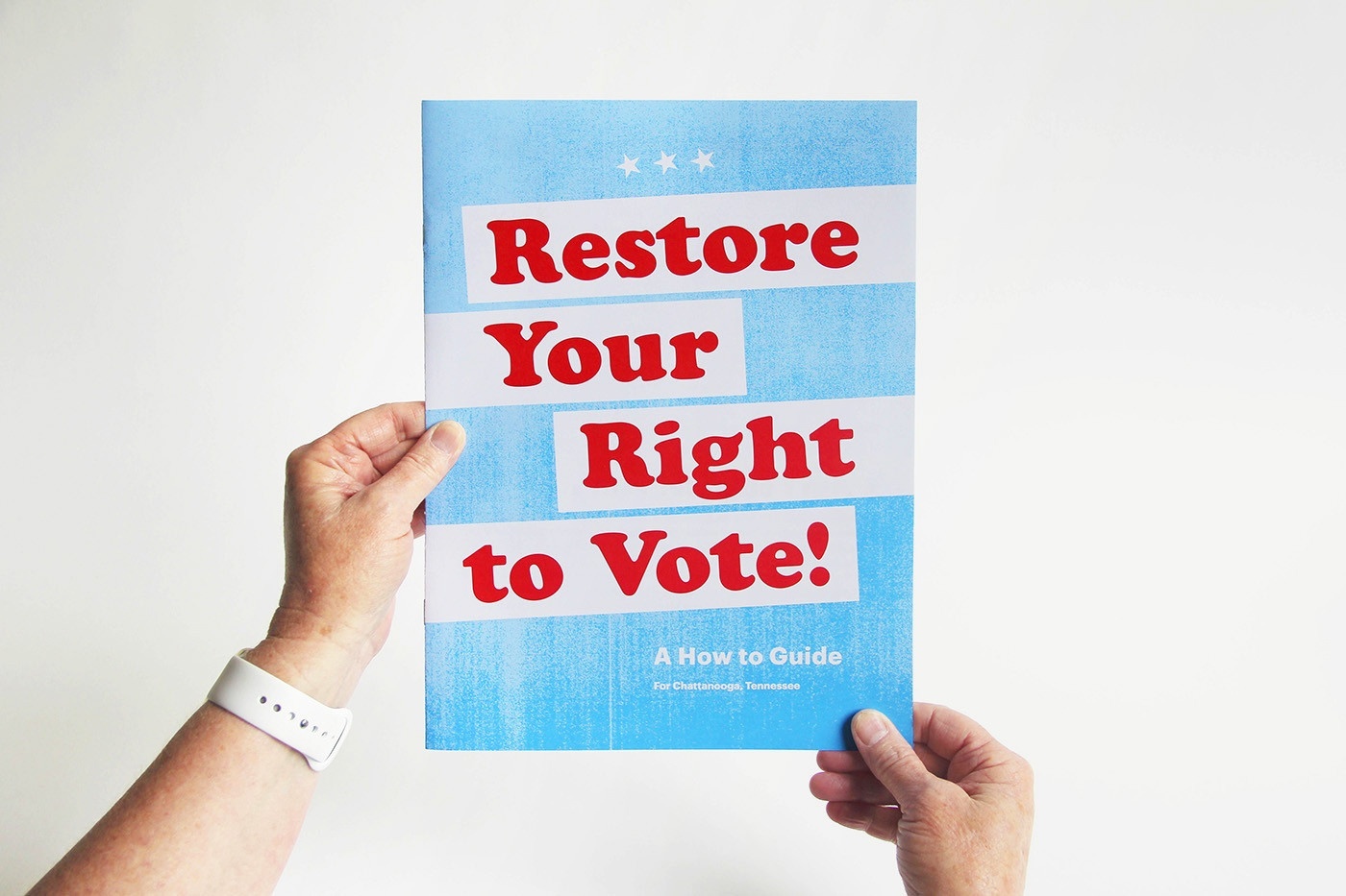 Photo of two hands holding the cover of a booklet that's sky blue, with white bands and bold red type that says Restore Your Right to Vote!