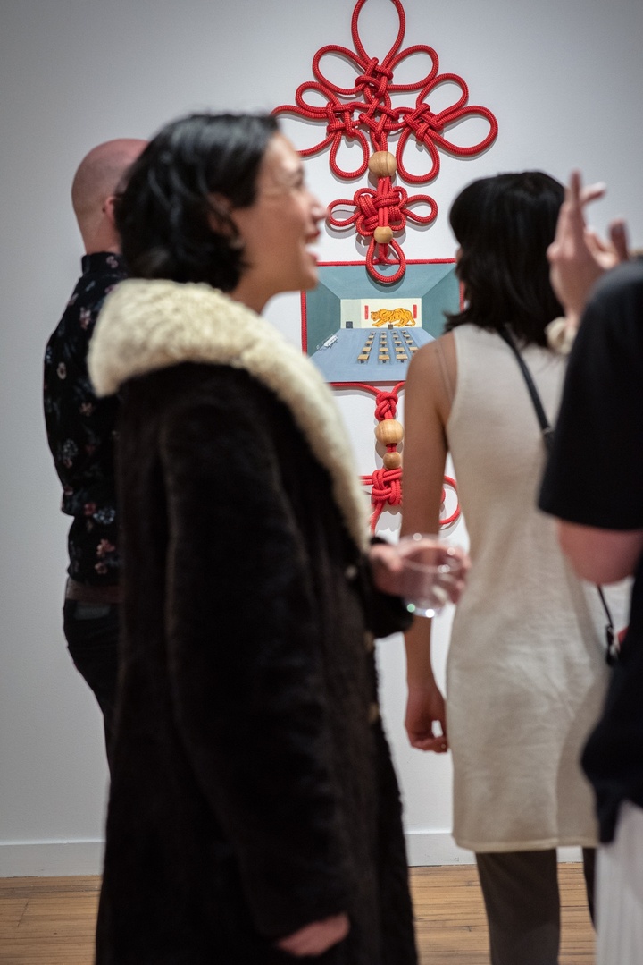 Several gallery visitors chat in the foreground in front of a piece of wall art with red decorative knotted rope framing a small painting of a meeting room.