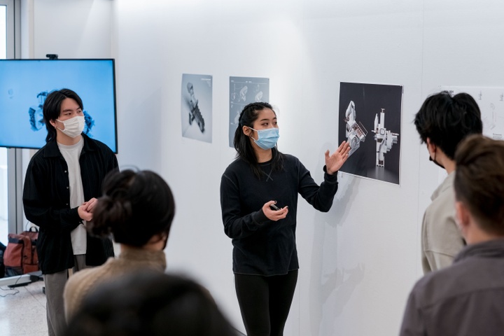 Two students stand in front of a group in the gallery space, discussing an architectural rendering hung on the wall.