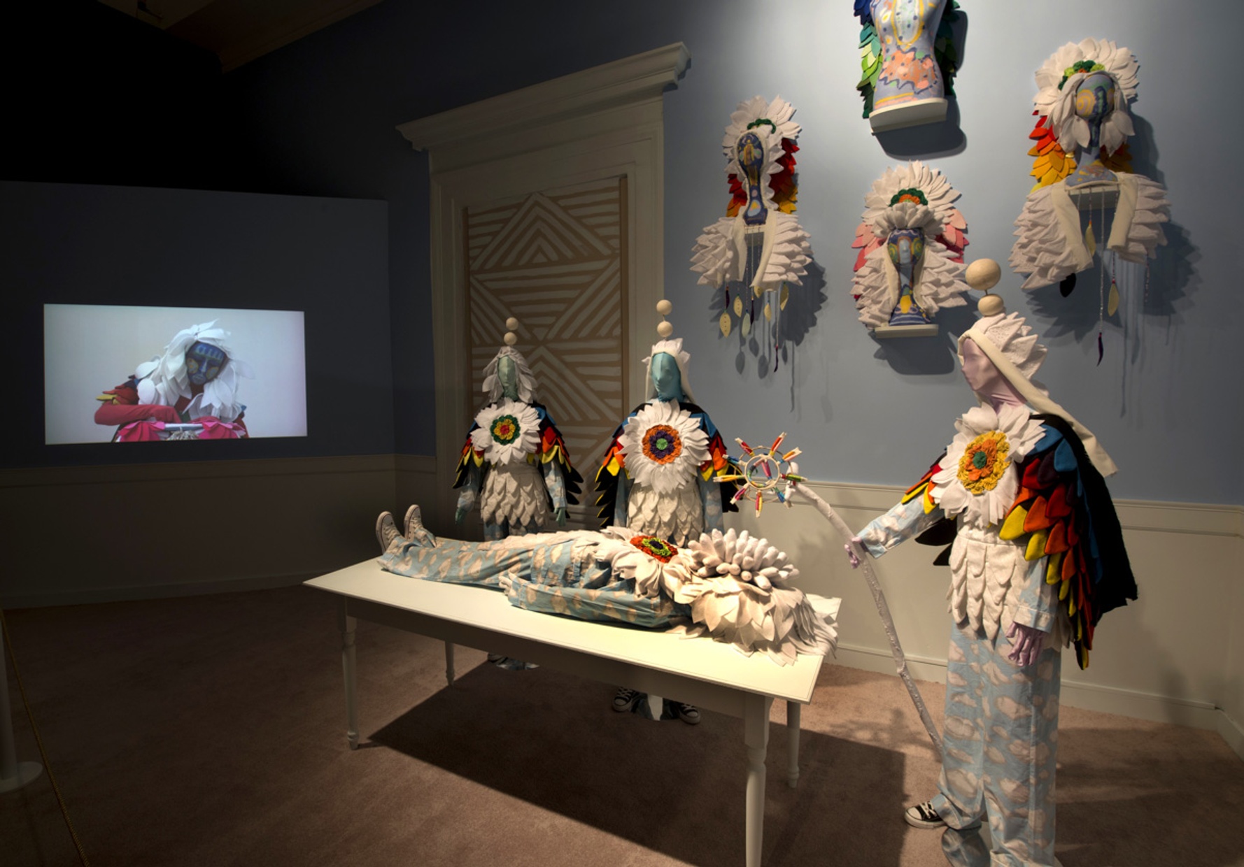 Three similarly dressed figures in blue, cloud pants and petal capes surround another figure lying on a table. Four, petal-like headdresses hang on a blue wall and a TV screen depicting another figure rests at the end of the table.