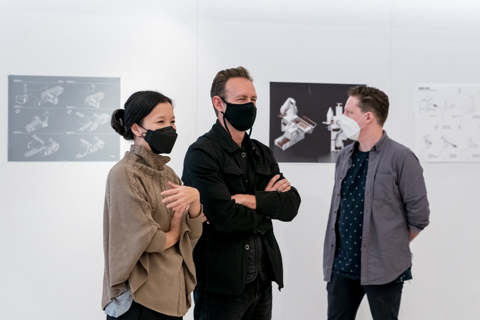 Two smiling people stand side by side in a gallery space in front of a a wall of architectural rendering prints.