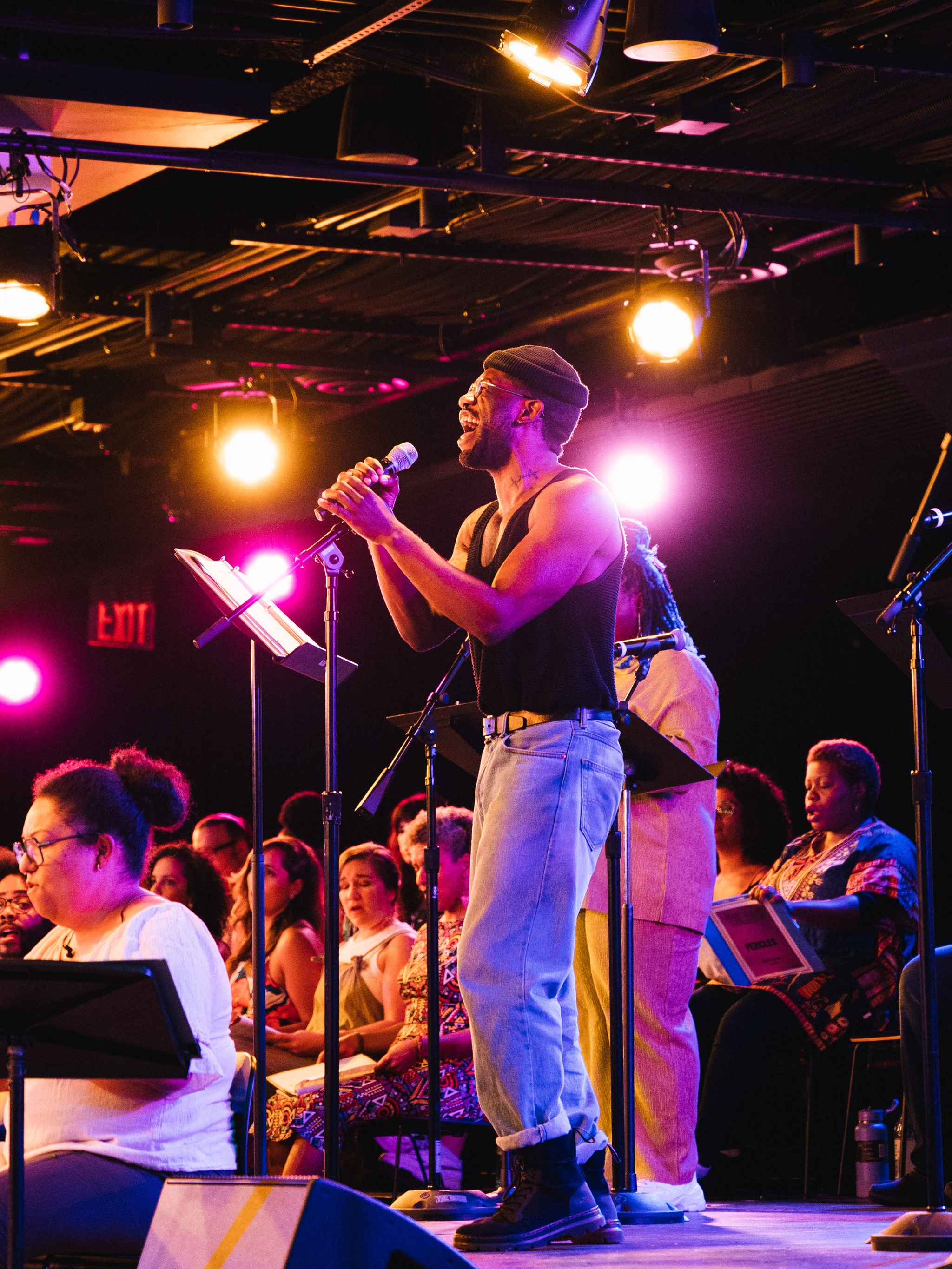 A Black man stands on a low stage at a microphone. He is holding it in both hands while he sings into it. He wears light blue jeans, a black tank top, and a black beanie.