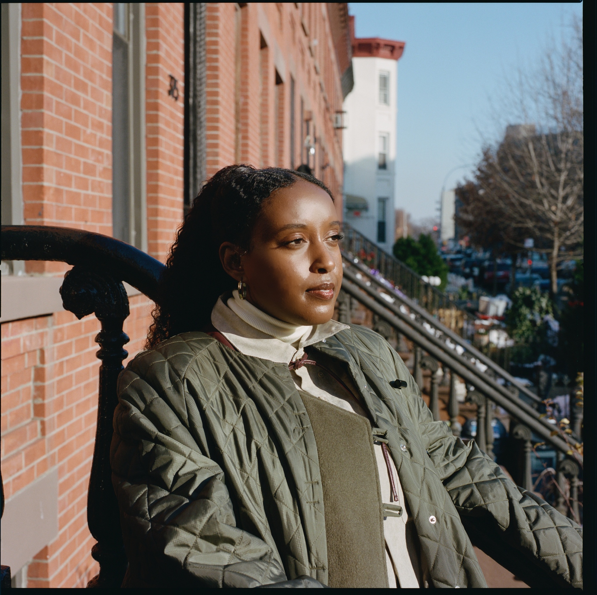 The artist Salome Asega in a green jacket looking out from a stoop against a line of red brick townhouses