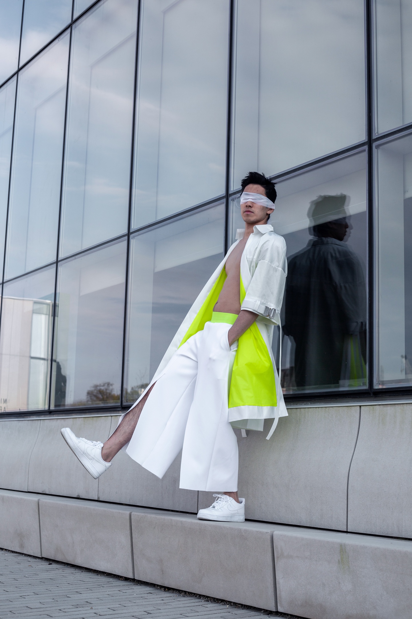Model wears a knee-length white overcoat with 3/4 sleeves and a neon green lining with white wide-leg pants with a neon green waistband, pockets, and slits in front. Model has a strip of white gauze tied across their eyes.
