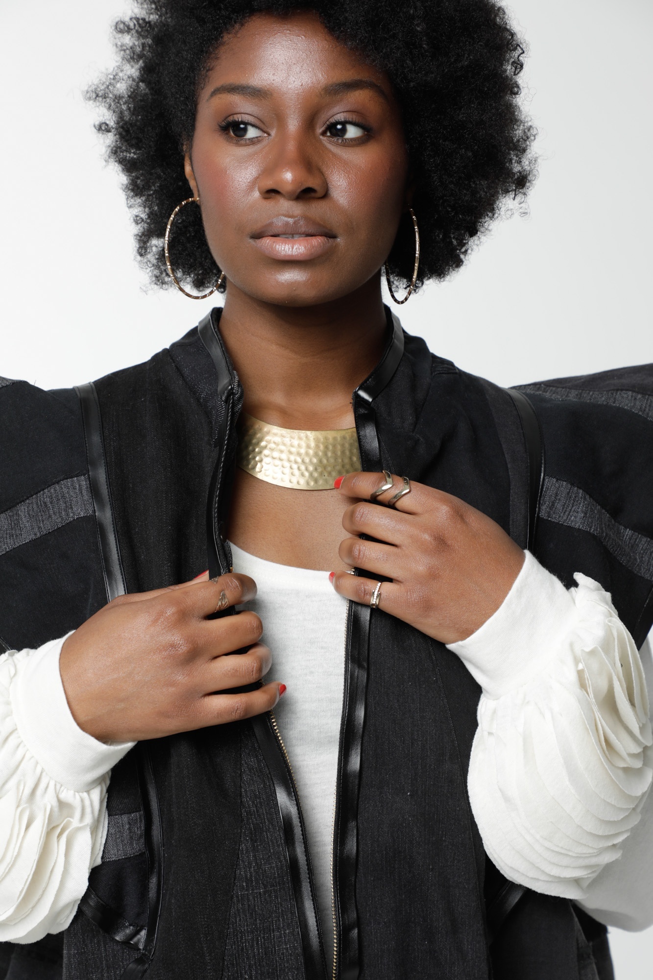 Closeup detail of a structured black jacket overlaid on a white long sleeved shirt. The jacket flares at the sleeve and the model wears a golden curved necklace.